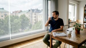 Mies istuu helsinkiläiskodin ikkunan ääressä, aurinkovarjon suojassa.